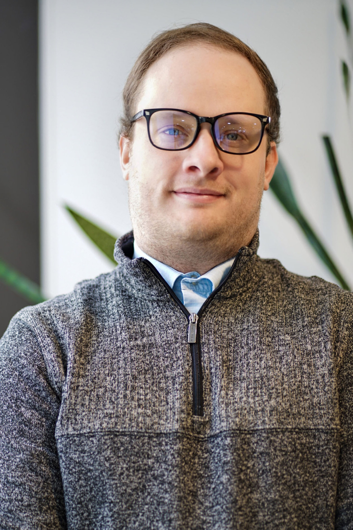 Headshot of Austin Haas smiling with short brown hair, black rimmed glasses, a brown quarter zip sweater and a light blue collared shirt