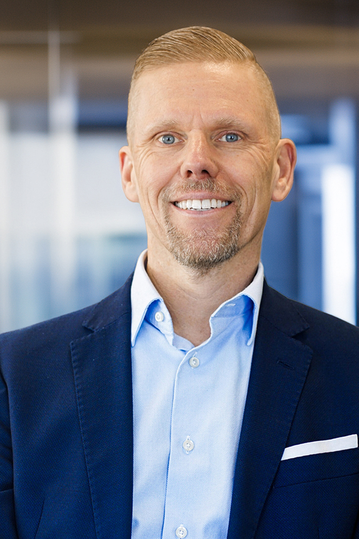 Headshot of John Burcke smiling with short blond hair, a dark blue blazer, and light blue collared shirt with no tie