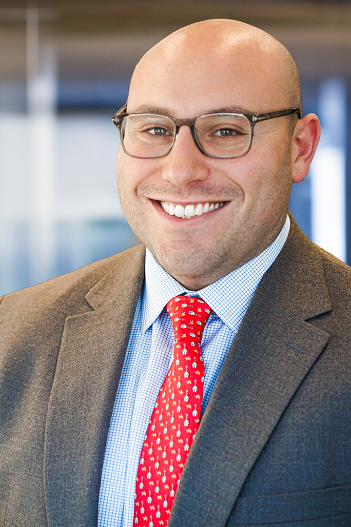 Headshot of Matt Lauro smiling with shaved head, brown glasses, brown blazer and blue and white checked shirt with a bright red patterned tie