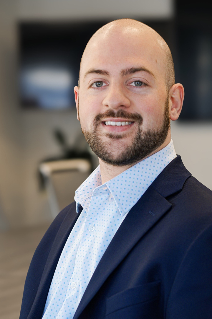Headshot of Matt Lambert smiling with shaved head, short beard, dark blue blazer, and a blue and white dotted shirt with no tie