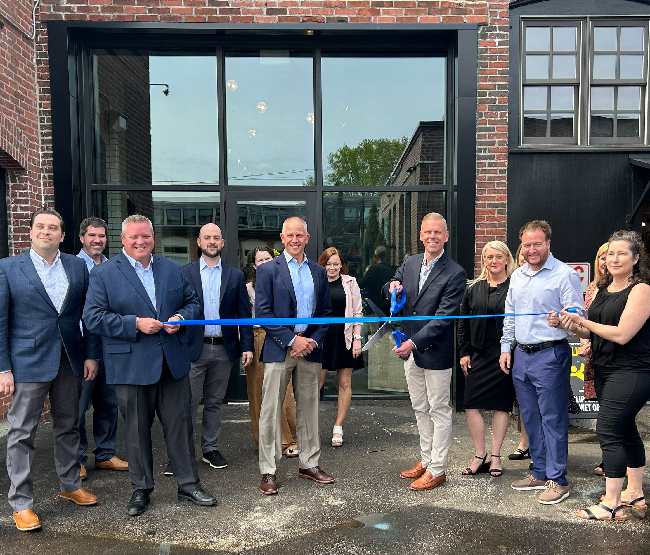 Group of smiling professional men and women celebrating the opening of the office by cutting a blue ribbon with giant scissors