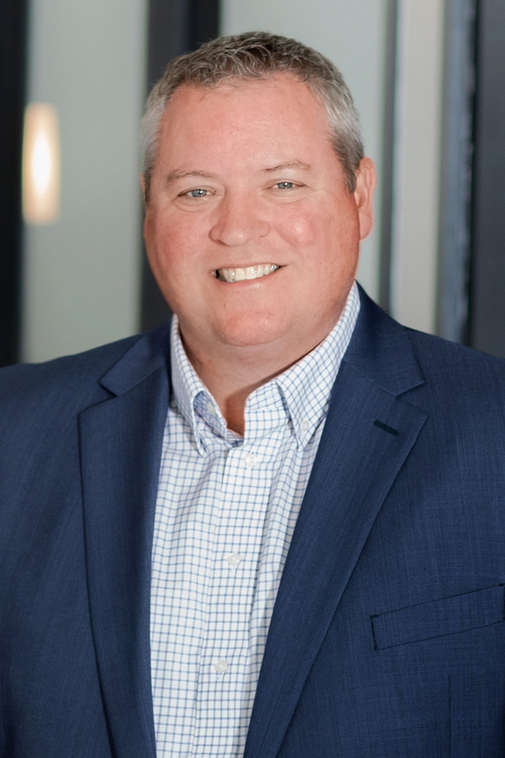 Headshot of Scott Eaton smiling and wearing a blue blazer with a blue and white plaid shirt and no tie