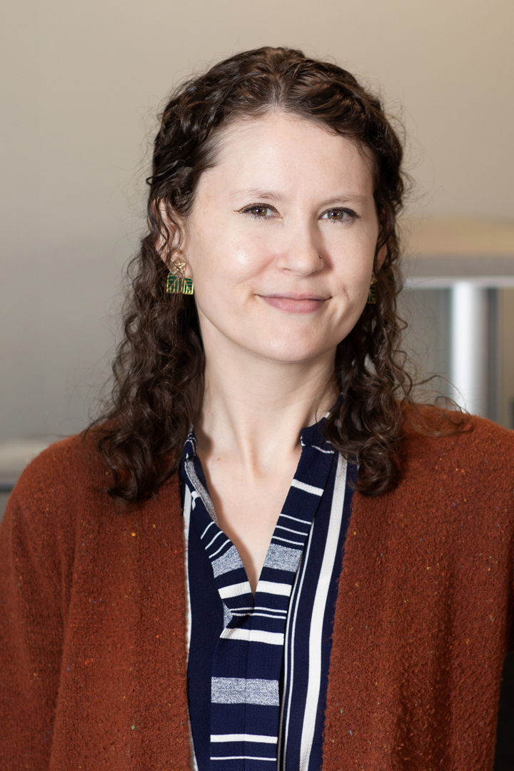Headshot of Stephanie Riggs smiling with long curly hair and a navy blue and white patterned blouse topped with a brown sweater